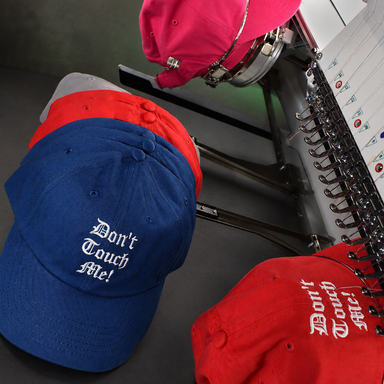 Blue, red, gray, and pink Don't Touch Me caps at an embroidery machine