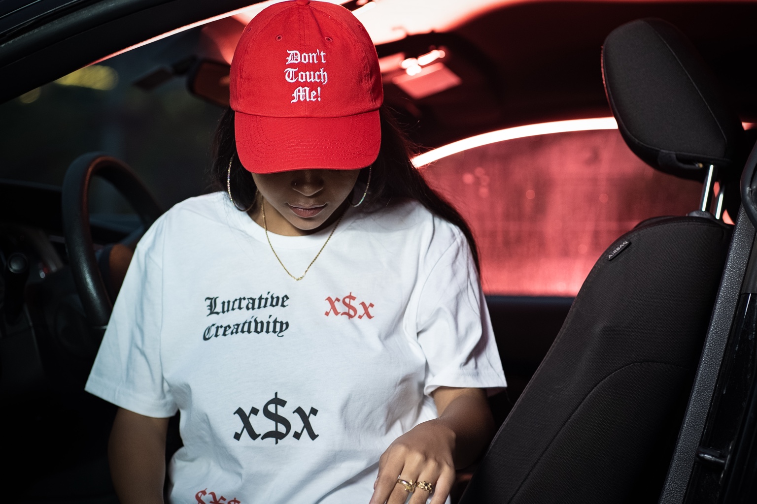 Woman wearing a red Don't Touch Me cap and Lucrative Creativity T-shirt inside a car